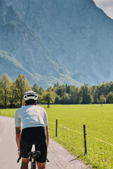 Cyclist riding on a paved road with mountains and a green field as the backdrop.  Active lifestyle and sports concept. Outdoors, back view. 