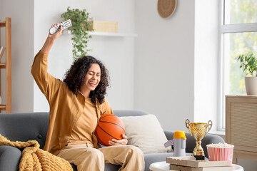 Female African-American sports fan with ball watching basketball on sofa at home