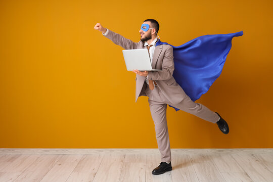 Young businessman dressed as superhero with laptop on orange background