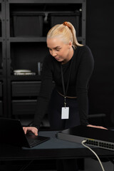 Female IT network technician in her twenties reviewing laptop beside network switch in server room