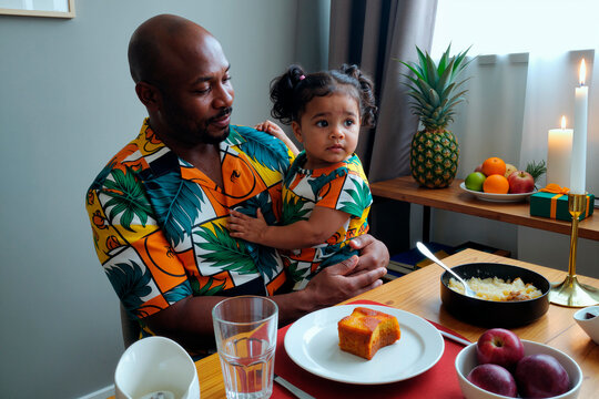 Black middle aged man holding young child with pigtails while sitting at dining table, sharing meal together, fruit and candles visible in background, both wearing matching shirts