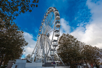 A giant white Ferris wheel rises against a dramatic blue sky with sweeping white clouds. Framed by dark-leafed trees and seen from a low angle, this iconic structure dominates the urban landscape