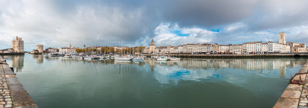 A panoramic view of La Rochelle's Vieux Port, featuring the iconic medieval defense towers and historic white buildings reflected in the still harbor water. Numerous yachts are docked under a dramatic - Powered by Adobe