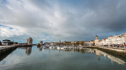 Fototapeta premium A panoramic view of La Rochelle's Vieux Port, featuring the iconic medieval defense towers and historic white buildings reflected in the still harbor water. Numerous yachts are docked under a dramatic