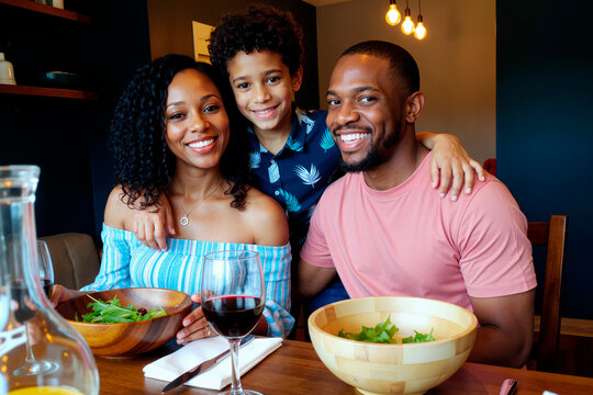 Black woman, Black man, and Black boy sitting together at dining table smiling, boy standing behind adults with arms around their shoulders, adults holding salad bowls, wine glass visible