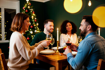 Group of young adult Caucasian and Black friends sitting at dining table clinking wine glasses, celebrating festive holiday together with Christmas tree and candles in background