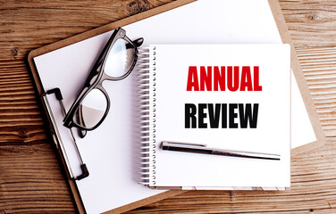 Spiral notebook with annual review text, pen and glasses on wooden office desk