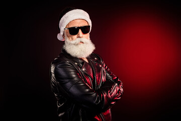 Cool Santa rocker in a leather jacket with white beard and dark sunglass standing against a red...