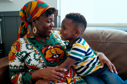 Black middle aged woman smiling and embracing boy sitting on couch, both looking at each other with joyful expressions, showing close family relationship and affection