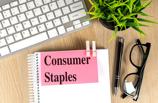 Words consumer staples written on pink note clipped to notebook with keyboard, pen, glasses and plant