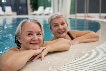 Two smiling senior women enjoying leisure time in an indoor swimming pool, leaning on the poolside. Concept of friendship, wellness, active lifestyle, relaxation, and healthy aging.