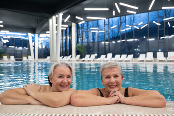 Two smiling senior women enjoying leisure time in an indoor swimming pool, leaning on the poolside. Concept of friendship, wellness, active lifestyle, relaxation, and healthy aging.