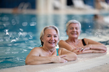 Two smiling senior women enjoying leisure time in an indoor swimming pool, leaning on the poolside. Concept of friendship, wellness, active lifestyle, relaxation, and healthy aging.
