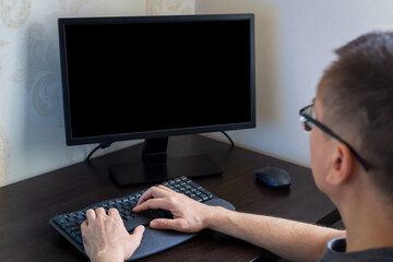 Remote work from home. Young man wearing glasses is focused on computer with black screen in stylish home office, featuring wooden desk and minimalistic decor, creating productive work environment