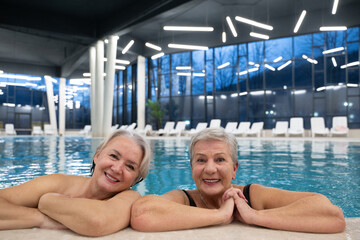 Two smiling senior women enjoying leisure time in an indoor swimming pool, leaning on the poolside. Concept of friendship, wellness, active lifestyle, relaxation, and healthy aging.