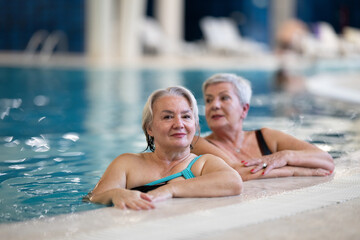 Two smiling senior women enjoying leisure time in an indoor swimming pool, leaning on the poolside. Concept of friendship, wellness, active lifestyle, relaxation, and healthy aging.