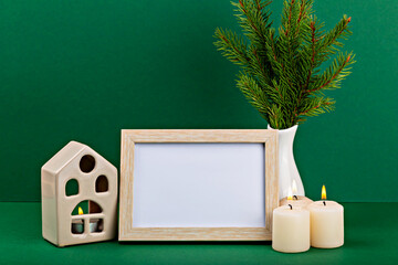 Mockup. Christmas. Decorative arrangement featuring an empty wooden frame, three lit candles, ceramic house, and an evergreen plant, creating cozy ambiance against vibrant green background
