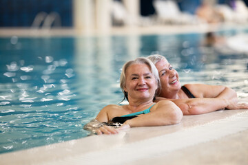 Two smiling senior women enjoying leisure time in an indoor swimming pool, leaning on the poolside. Concept of friendship, wellness, active lifestyle, relaxation, and healthy aging.
