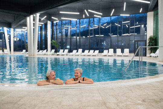 Two smiling senior women enjoying leisure time in an indoor swimming pool, leaning on the poolside. Concept of friendship, wellness, active lifestyle, relaxation, and healthy aging.