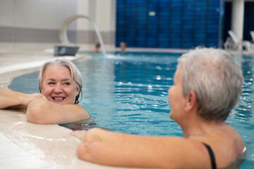 Fototapeta premium Two smiling senior women enjoying leisure time in an indoor swimming pool, leaning on the poolside. Concept of friendship, wellness, active lifestyle, relaxation, and healthy aging.