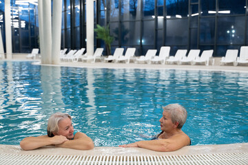 Two smiling senior women enjoying leisure time in an indoor swimming pool, leaning on the poolside. Concept of friendship, wellness, active lifestyle, relaxation, and healthy aging.
