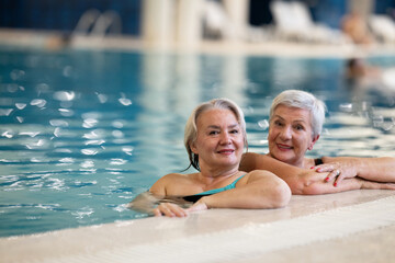 Two smiling senior women enjoying leisure time in an indoor swimming pool, leaning on the poolside. Concept of friendship, wellness, active lifestyle, relaxation, and healthy aging.