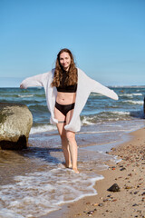 Young woman in black swimwear and white cover-up walking on Baltic Sea beach on sunny day