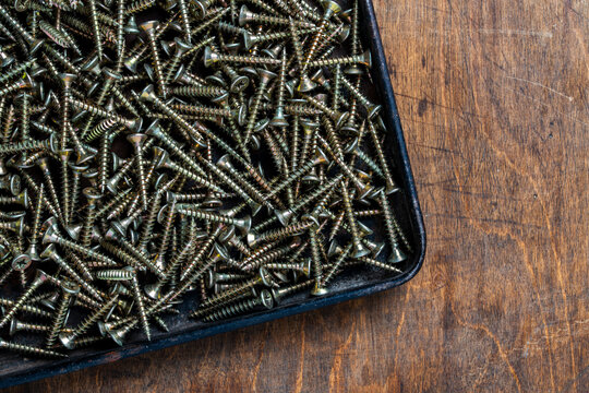 Many golden metal self tapping screws for construction work in black metal tray on wooden background , closeup, top view, copy space. New fasteners self tapping screws for connecting wooden elements