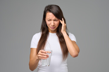 Woman with headache holding glass of water expressing pain or discomfort against gray background
