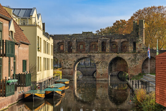 Historic Berkelpoort water gate in Zutphen, Netherlands, featuring medieval brick arches over the tranquil Berkel river lined with classic Dutch houses and narrow boats on a sunny autumn day.