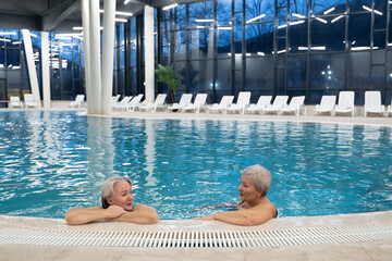 Two smiling senior women enjoying leisure time in an indoor swimming pool, leaning on the poolside. Concept of friendship, wellness, active lifestyle, relaxation, and healthy aging.