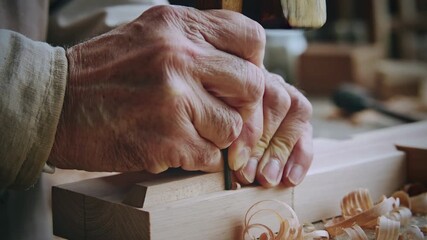 Skilled senior carpenter's wrinkled hands carving a piece of wood with a chisel, demonstrating traditional craftsmanship and precision woodworking in his workshop with shavings on the workbench