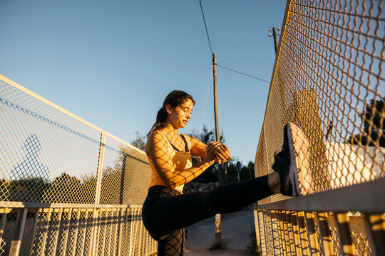Young woman stretching leg on fence before running workout