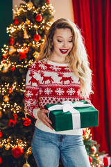 Funny christmas surprise as a smiling woman in a red and white sweater holds agreen wrapped gift by...