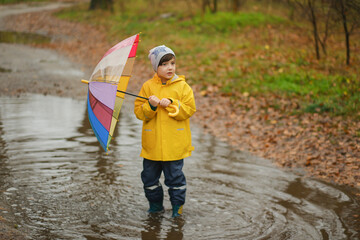 Happy child in yellow raincoat and hat holding colorful rainbow umbrella standing in puddle on rainy autumn day in park.