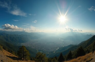 Bright sun shines over green mountain range and valley city. Hazy atmosphere covers town nestled between rolling hills. Serene natural landscape panorama shows vast urban sprawl below.