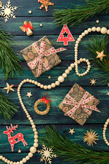 Fototapeta premium Christmas gifts with pinecone wrapping, red ribbon, pine branches, reindeer, angel, wreath, and festive ornaments on dark teal wooden surface.