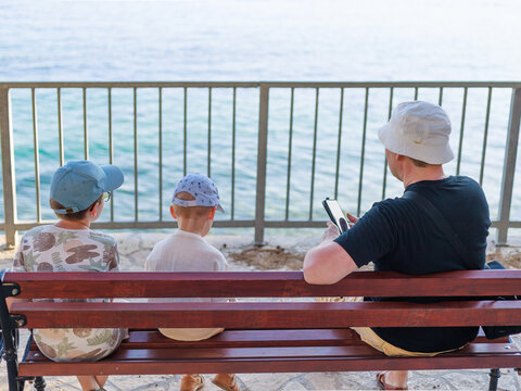 Man and two boys sitting on wooden bench facing the sea on summer day, copy space. Concept of family travel, relaxation, connection, peaceful vacation moment.