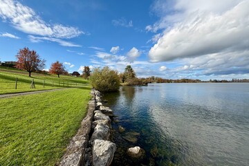 On the shore of Lake Zug in Buonas, Risch, a beautiful autumn day unfolds with crisp air, gentle waves, and a blue sky scattered with soft clouds.
