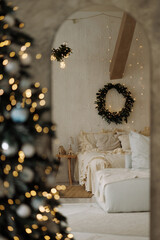 Warm bedroom with Christmas wreath on beige wall and blurred decorated tree in front. Concept of winter holidays, cozy home, festive interior, peaceful atmosphere.