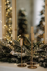 Elegant golden candle holders on decorated mantel with fir branches, fairy lights and mirror reflection. Warm bokeh background. Concept of Christmas, luxury decor, winter holiday, celebration.
