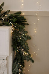 Evergreen Christmas garland with pearl and gold ornaments decorating white fireplace against beige wall. Warm fairy lights create festive atmosphere. Concept for holiday decor, winter, celebration.