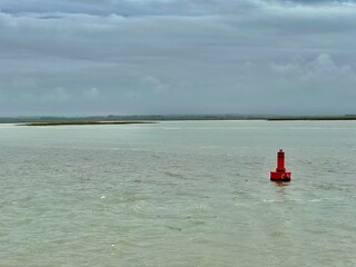 Red navigation buoy floating on the calm tidal waters of the Baie de Somme near Le Crotoy, France, under a dramatic overcast sky, with distant sandbanks and wetlands shaping a wide, maritime horizon