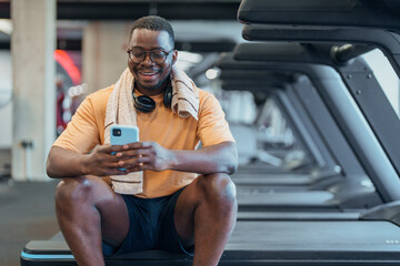 Smiling middle aged Black man resting with phone on gym bench with bright indoor lighting and towel
