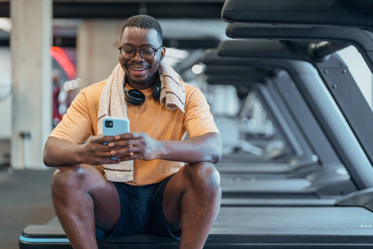 Smiling middle aged Black man resting with phone on gym bench with bright indoor lighting and towel