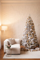 Christmas interior of a bright modern living room. In keeping with the 2026 New Year's trend. A snow-covered Christmas tree with beige and gold decor next to a white sofa against a textured wall.