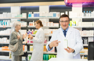 Obraz premium Polite middle-aged male pharmacist demonstrating preparation in box in chemist's shop with large assortment