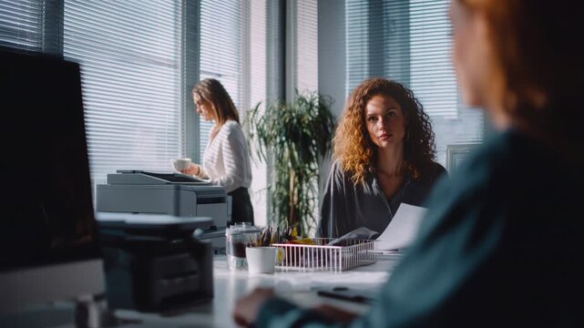 Three young businesswomen focusing on their tasks in a bright, modern office, with one colleague using a printer while another reviews documents and talks with a coworker at her desk