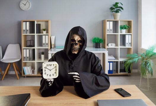 Portrait of overworked tired funny business man company employee in death grim reaper costume pointing his finger to the alarm clock sitting at workplace in office. Overload deadline concept.