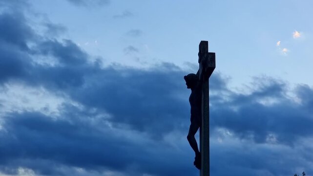Stone statue representing Jesus Christ silhouette on a cross with dramatic sunset sky, 4K clouds time lapse